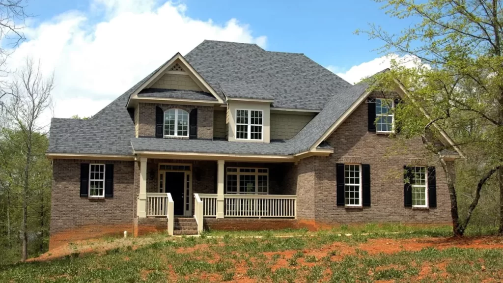 A new, two-story brown brick house with a gray roof and black shutters, situated on a sloped, unfinished yard of red dirt under a blue sky.