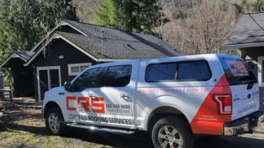 A white CRS Certified Roofing Services truck, a roofing company in Portland, is parked in front of a dark-colored house with a newly installed black shingle roof.