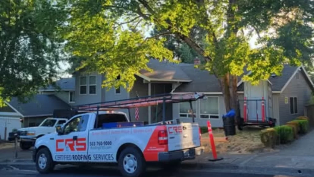 A work truck for CRS Certified Roofing Services, a roofing company in Portland, is parked on a street in front of a residential home with a large, leafy green tree.