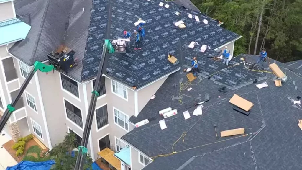 Roofing contractors installing shingles and underlayment on a large multi-story building using safety harnesses and cranes, demonstrating professional roofing work in Portland.