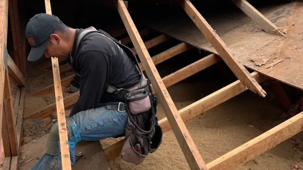 A roofer in full gear working on repairing the damaged wooden frame of a roof from inside an attic space.